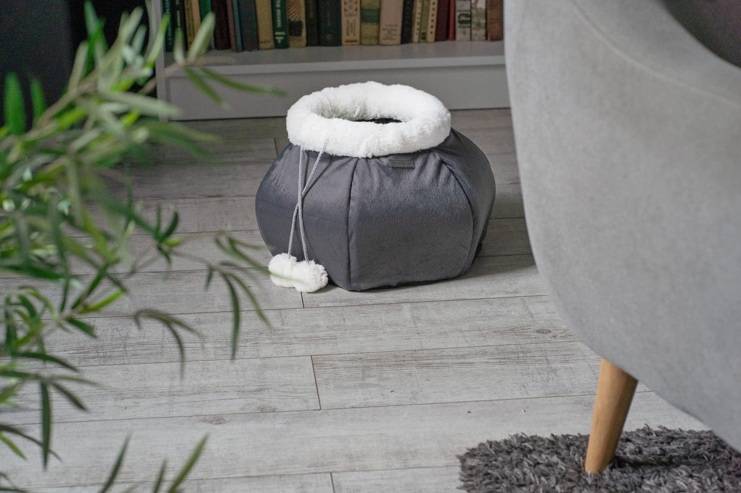 Gray pet bed with white pom-poms on a wooden floor next to a gray armchair.