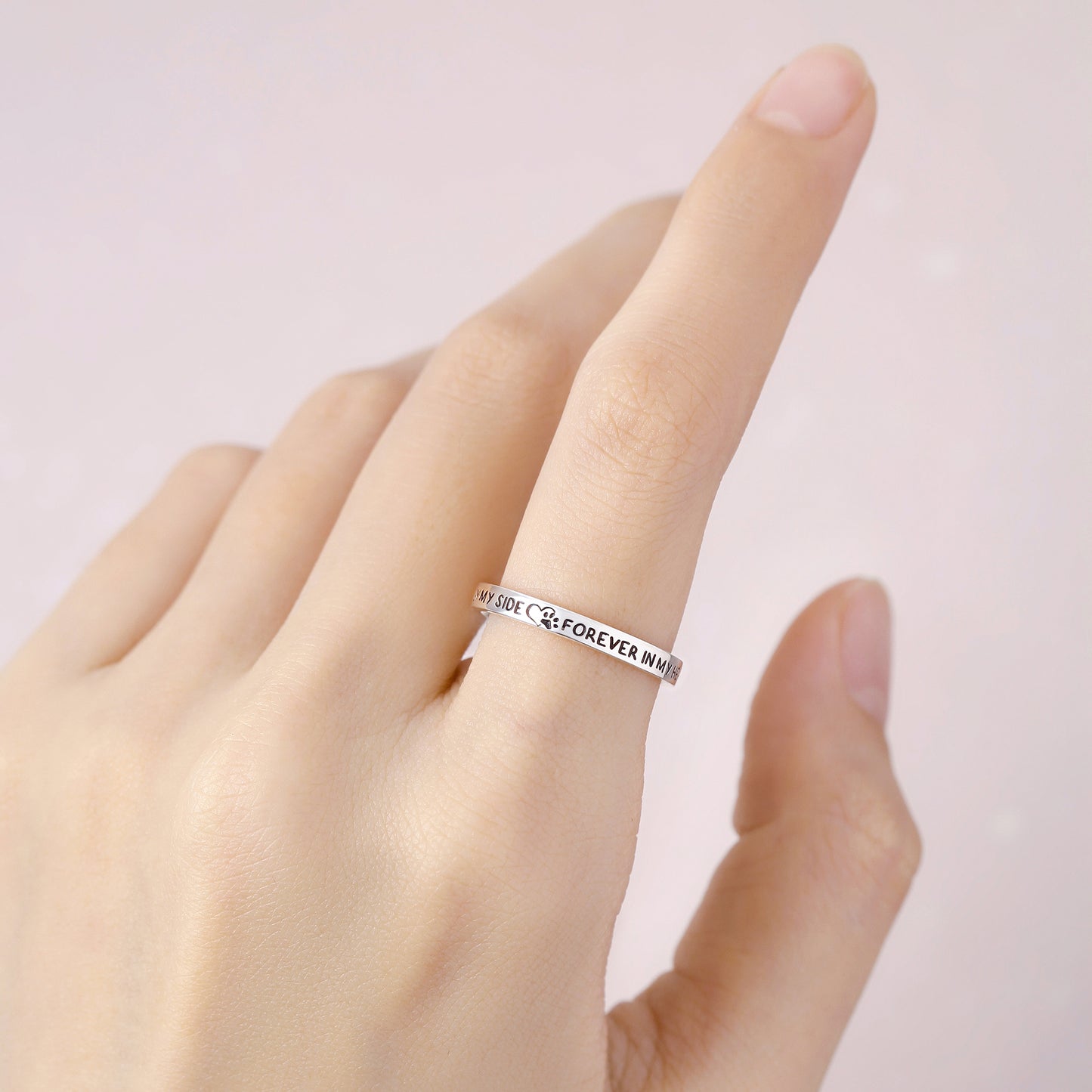 Silver ring with engraved text on a hand against a light pink background