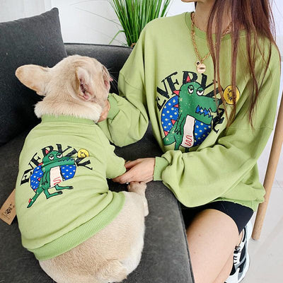 Person and dog wearing matching green sweatshirts with a cartoon dinosaur design.