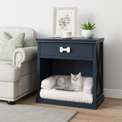 Navy blue pet bed with a cat inside, placed on a wooden floor next to a gray armchair.