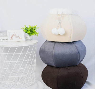 Stack of three round cushions in beige, gray, and brown on a white surface with a small plant and books in the background.
